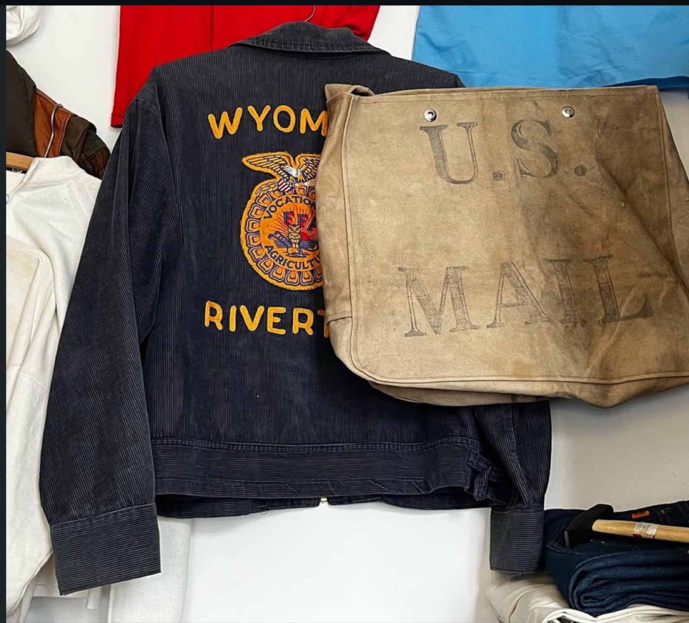 Black corduroy jacket embroidered Wyoming Riverton with a Future Farmers of America emblem, hanging beside a tan canvas U.S. Mail carrier bag